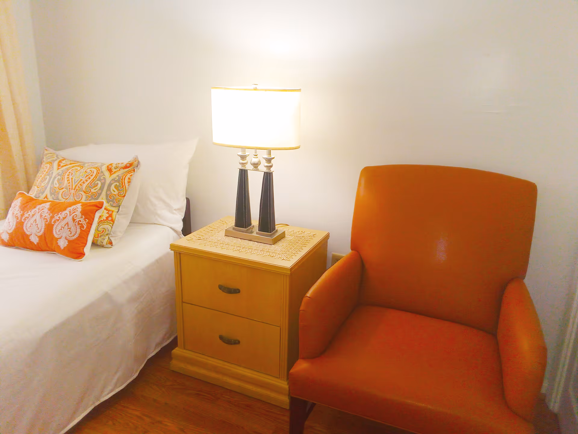 A cozy corner of a bedroom featuring a bed with white sheets and decorative pillows in orange and patterned designs, a wooden nightstand with two drawers, and a table lamp with a white shade. Next to the nightstand is an orange upholstered armchair on a wooden floor.