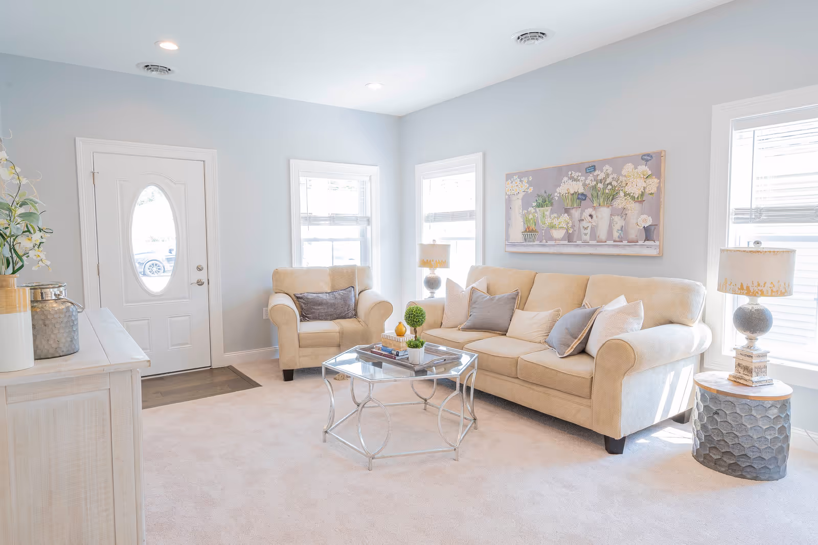 A bright and cozy living room with a beige sofa and armchair, both adorned with multiple cushions. A glass coffee table with decorative items sits in the center on a light carpet. The room has light blue walls, two windows with blinds, a white front door with an oval window, and a side table with a lamp. A floral painting hangs on the wall above the sofa.