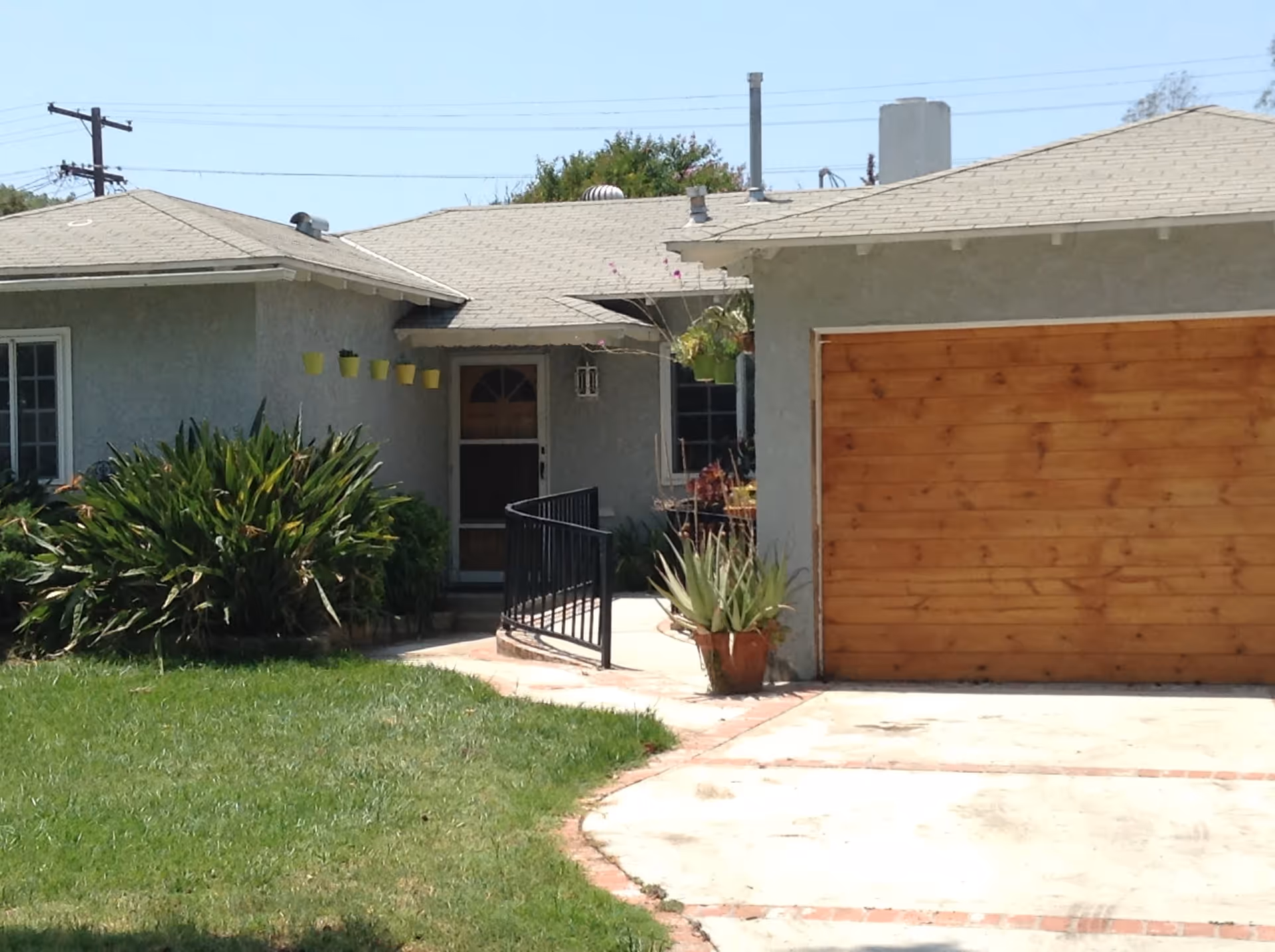 Exterior view of a single-story residential building with a gray roof and walls, a wooden garage door, a concrete driveway, a green lawn, and various plants near the entrance. There is a black metal handrail along a small ramp leading to the front door.