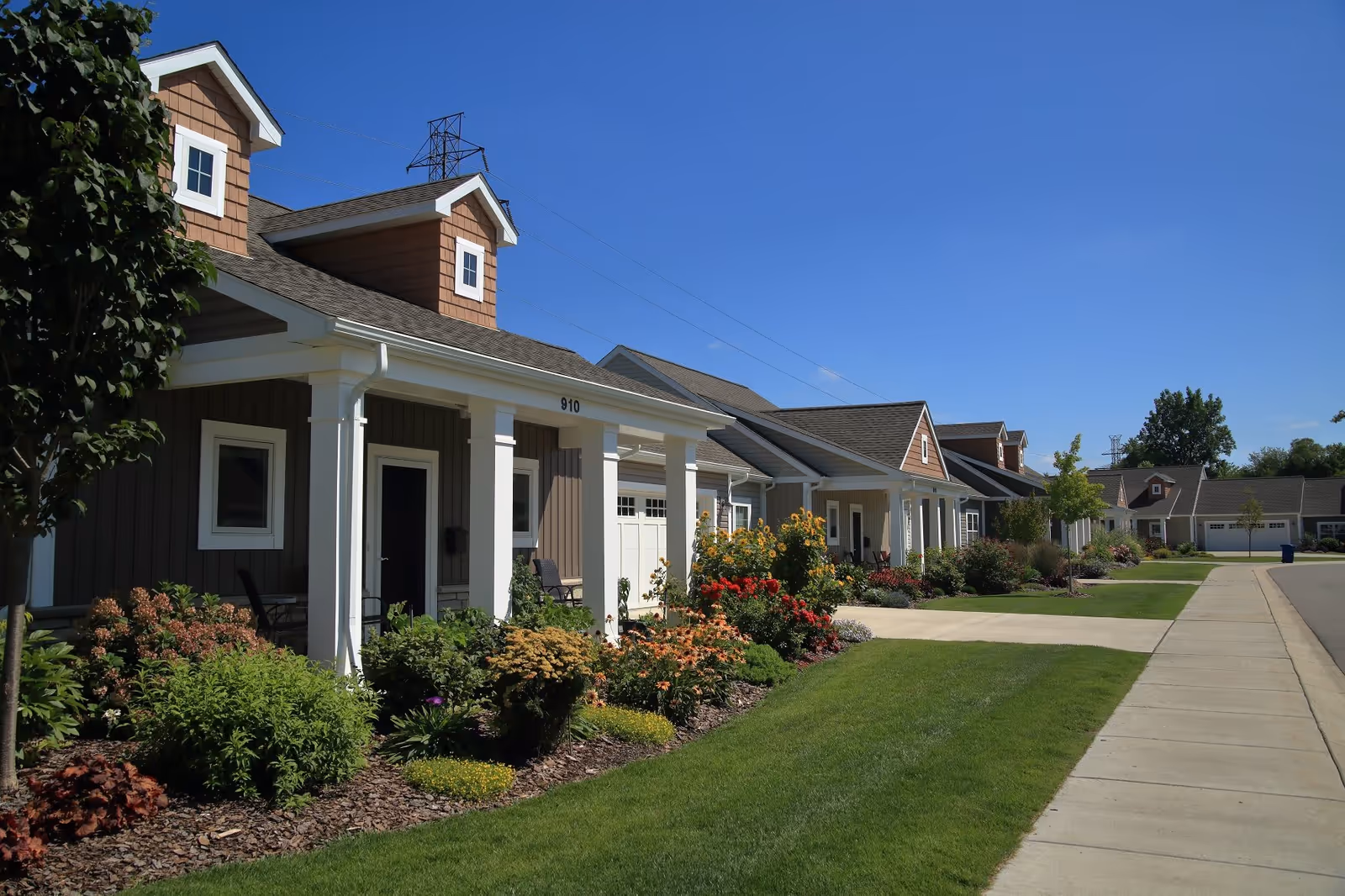 Row of single-story residential buildings with porches, surrounded by well-maintained gardens and green lawns under a clear blue sky.