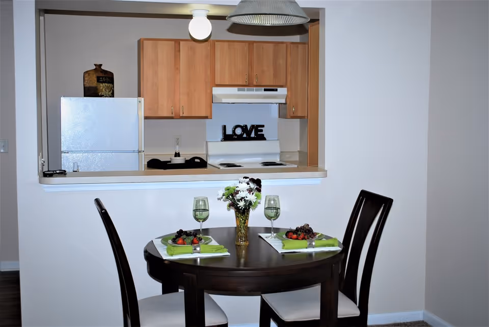Small dining area with a round dark-wood table set for two and a pass-through view into a kitchen with wooden cabinets and a 'LOVE' sign.