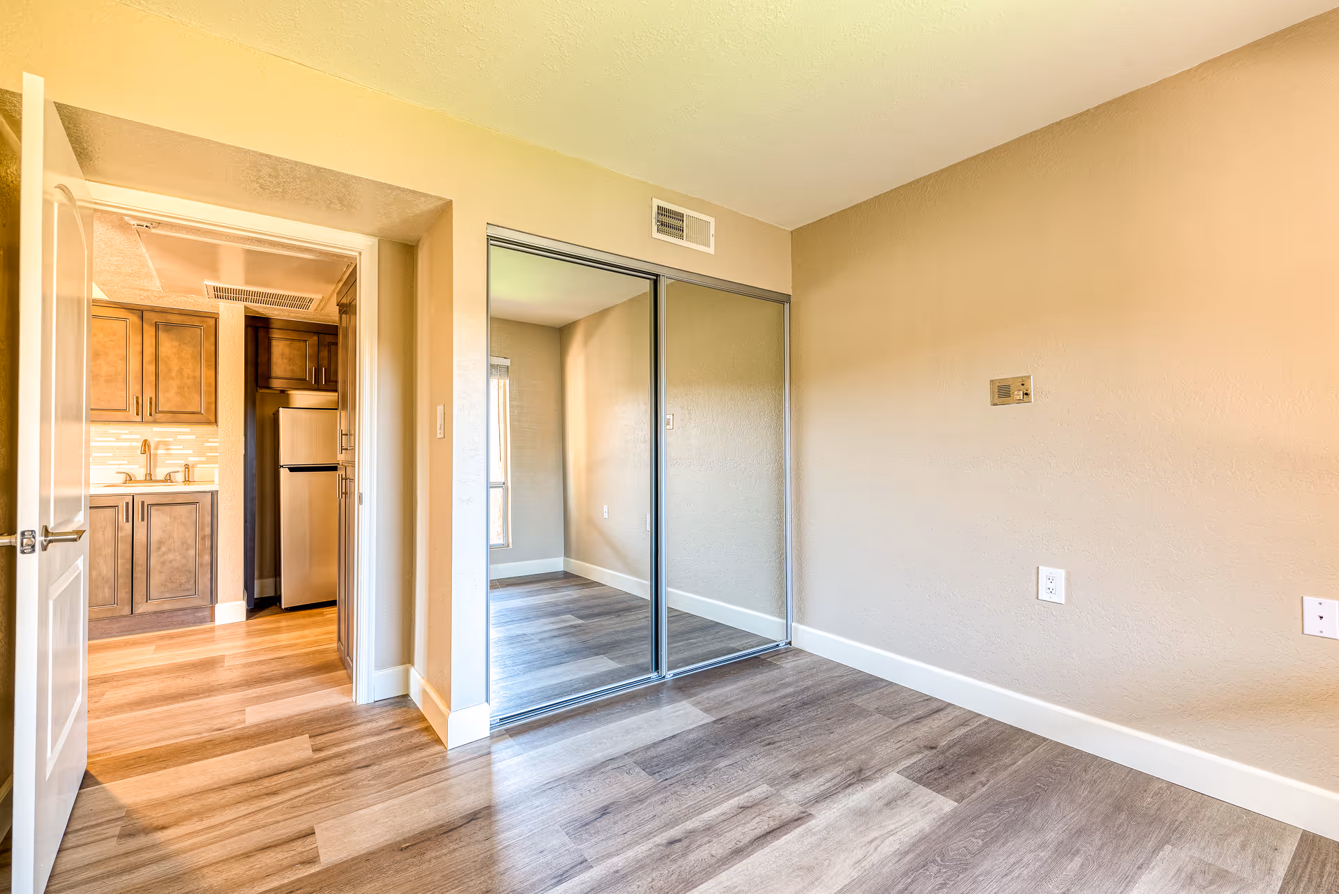 Empty bedroom with mirrored sliding closet doors, wood-look flooring, and a view into a kitchen area.