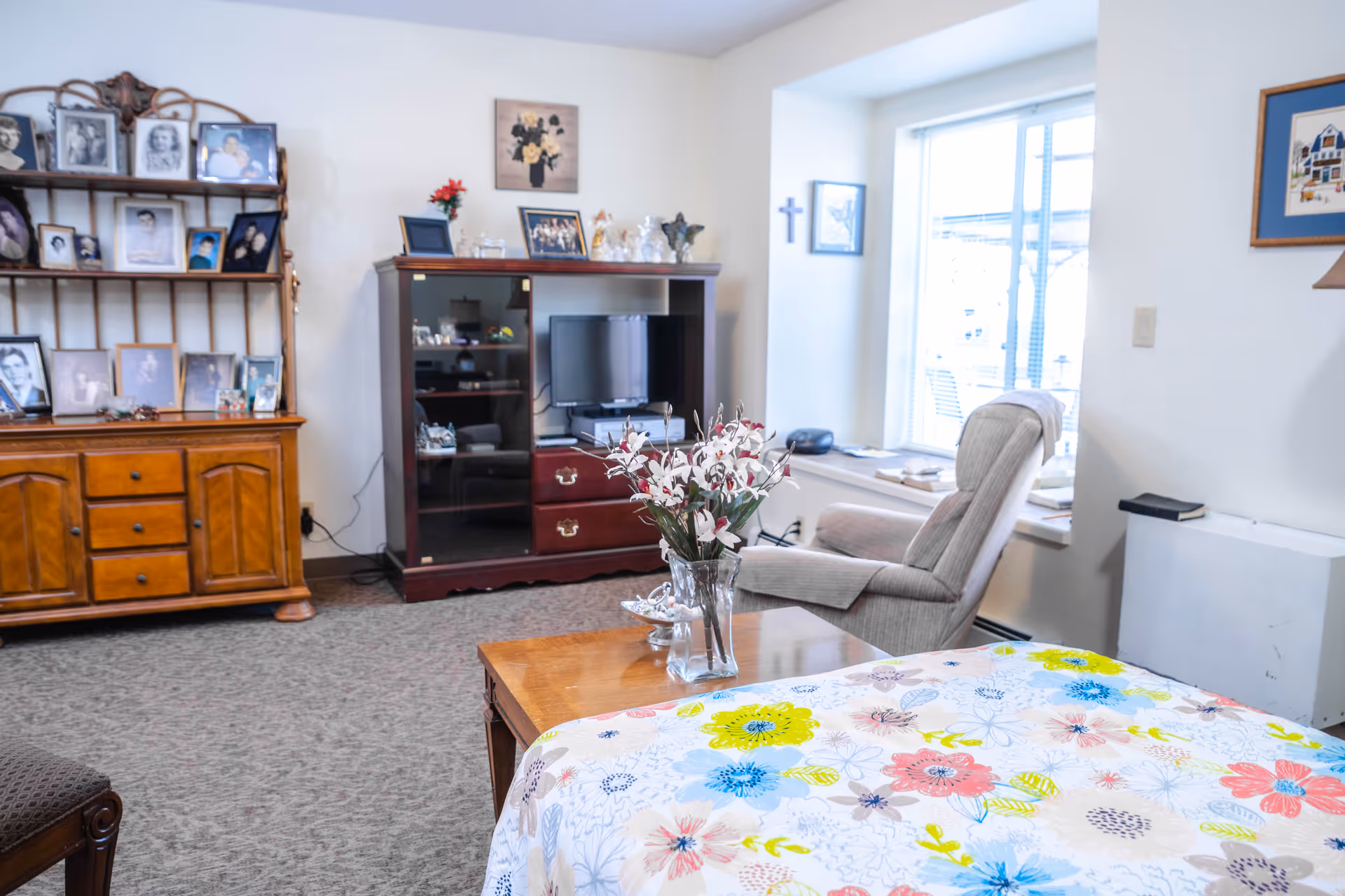 A cozy living room area with a wooden cabinet filled with framed photos on the left, a dark wood entertainment center with a TV in the middle, and a comfortable recliner chair near a window on the right. A wooden coffee table with a vase of flowers is in the foreground, and a colorful floral blanket is partially visible.