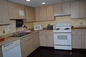 Interior view of a kitchen with light wood cabinets, a white stove with oven, a microwave, a toaster, a dishwasher, and a stainless steel sink. The countertops are beige, and the floor is dark-colored.