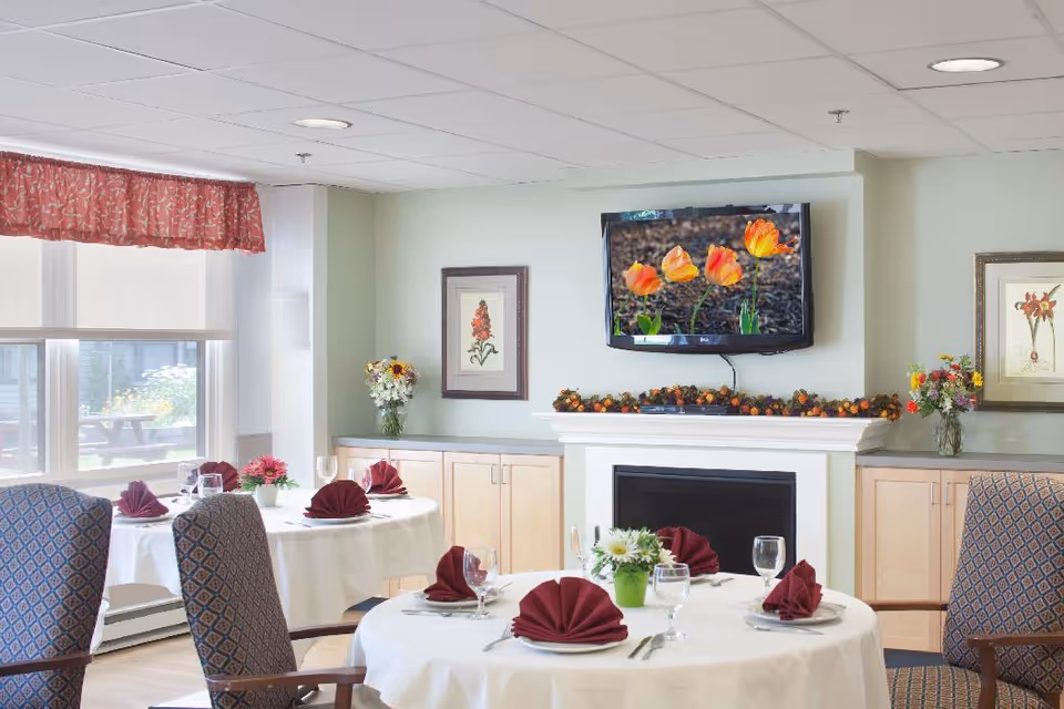 A dining room with round tables covered in white tablecloths, each set with plates, glasses, silverware, and burgundy folded napkins. There are patterned upholstered chairs around the tables. A fireplace with a garland on the mantel and a flat-screen TV displaying orange flowers is mounted on the wall. The room has large windows with red valances, framed floral artwork, and flower arrangements on the mantel and tables.