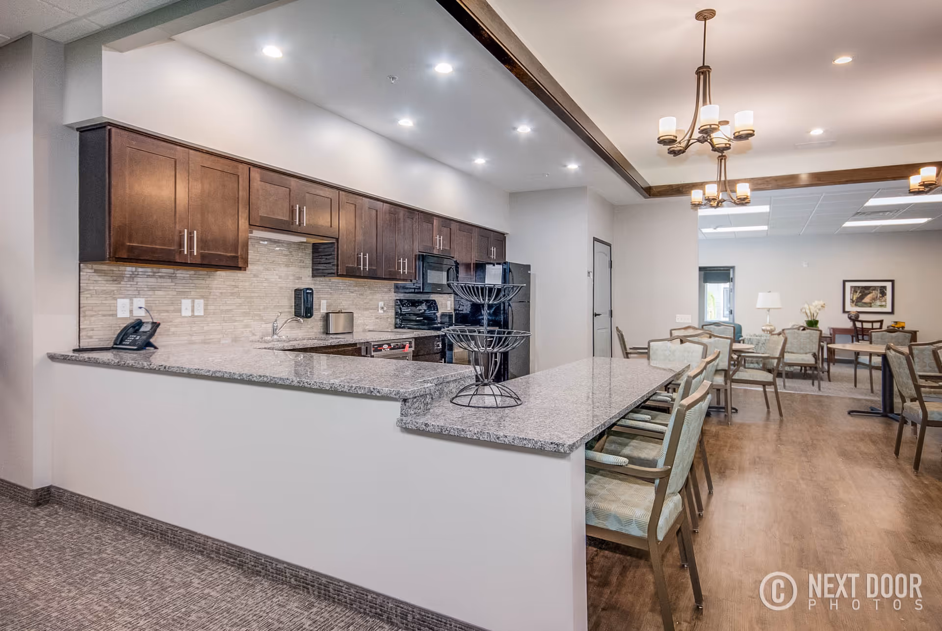 Open communal kitchen and dining area with a long granite island, wooden cabinets, multiple chairs and tables, and chandeliers.