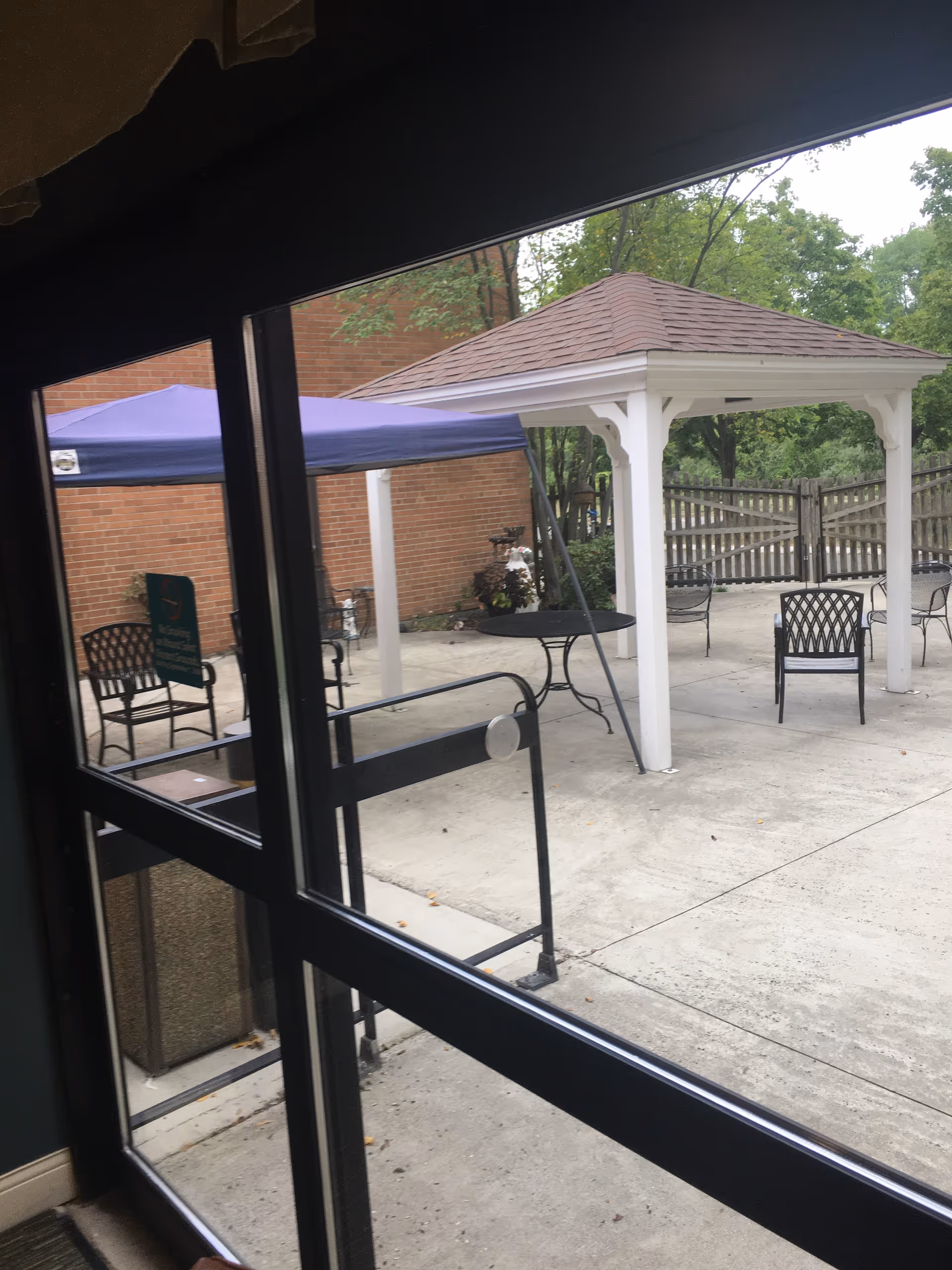 View through glass doors of an outdoor courtyard with a white gazebo, metal chairs and tables on a concrete patio.