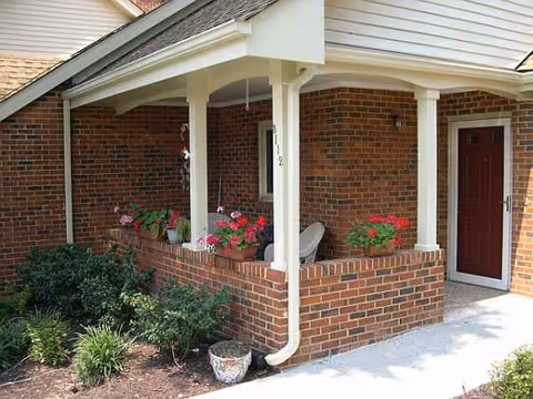 Brick exterior porch with white columns, potted flowers, and a red front door.