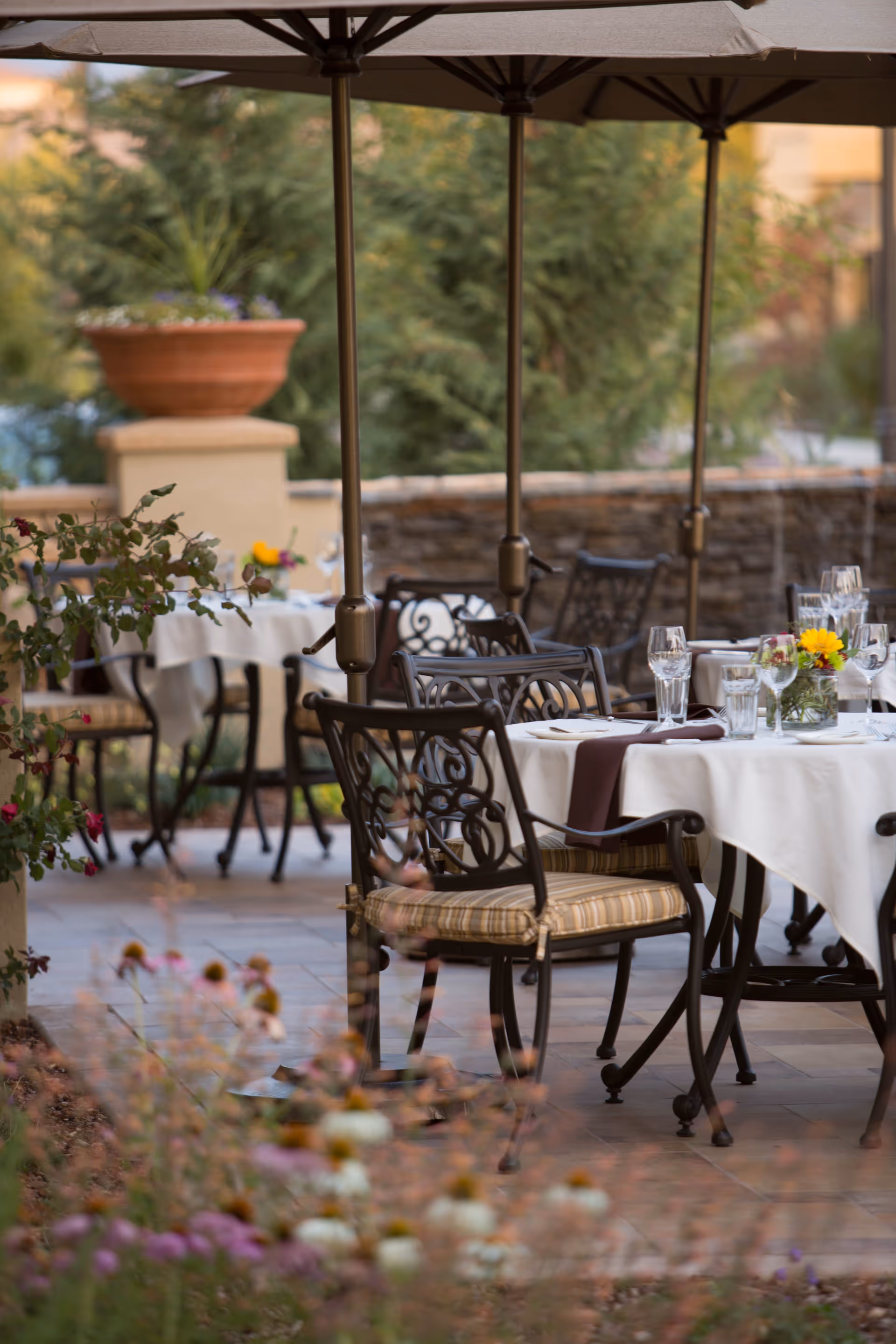 Outdoor patio dining area with tables covered in white tablecloths, set with glassware and floral centerpieces. Ornate metal chairs with striped cushions surround the tables, and large umbrellas provide shade. The background includes greenery and a stone wall.