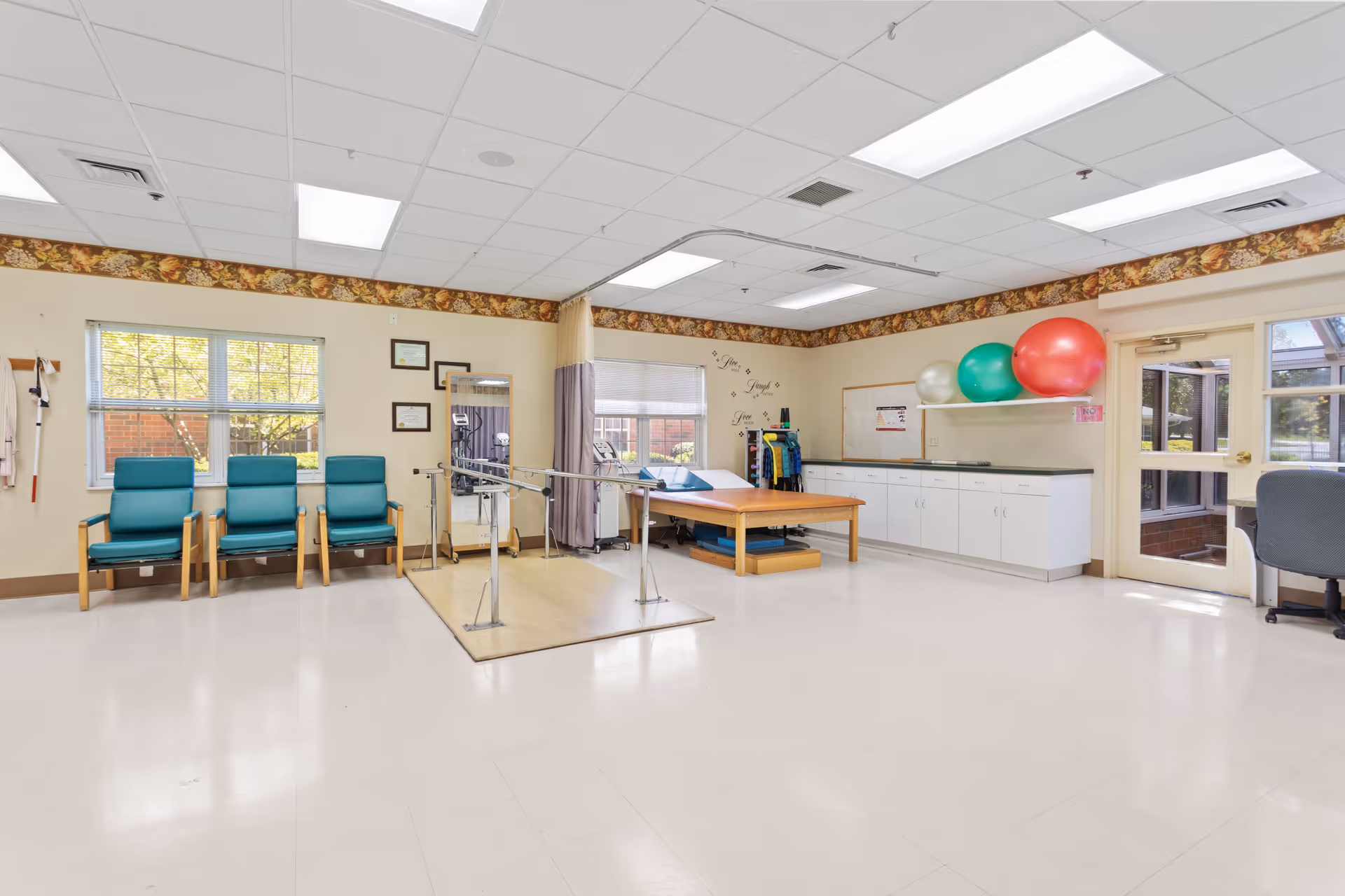A spacious rehabilitation room with three teal chairs lined up against a wall under two windows. In the center, there are parallel bars for walking exercises and a padded therapy table. On the right side, there are white cabinets with three large exercise balls on top and a door leading outside. The room has a white tiled floor, a drop ceiling with fluorescent lights, and a floral border along the top of the walls.