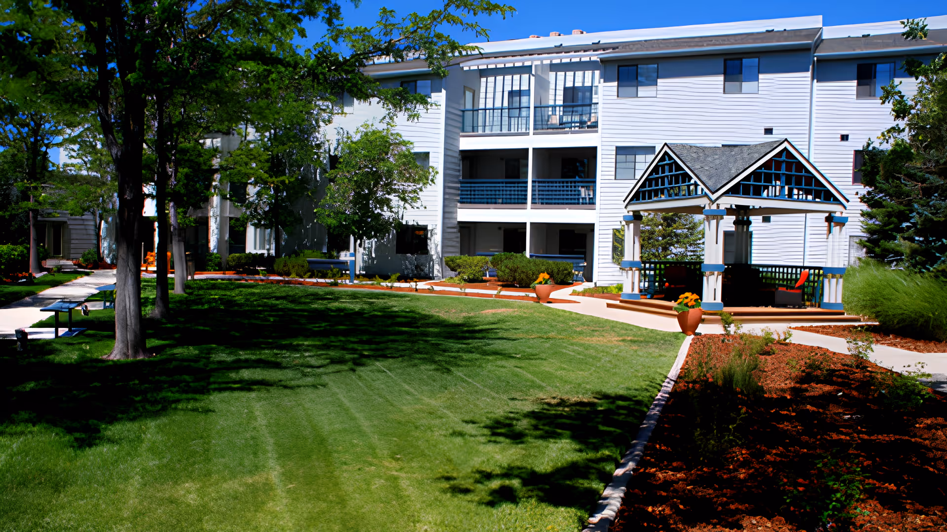 Outdoor view of a senior living facility with a well-maintained lawn, trees, a paved walkway, benches, and a gazebo. The building in the background is white with multiple windows and balconies.