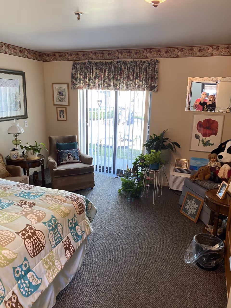 A cozy bedroom with a bed covered in an owl-patterned quilt, a cushioned armchair with a decorative pillow, several potted plants near a sliding glass door with vertical blinds, framed artwork on the walls, a mirror reflecting two people taking a photo, and a small table with a lamp and other decorative items.