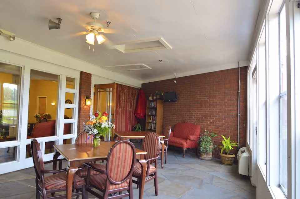Sunlit communal dining and sitting area with a wooden table and striped chairs, a red armchair against a brick wall, and large windows.