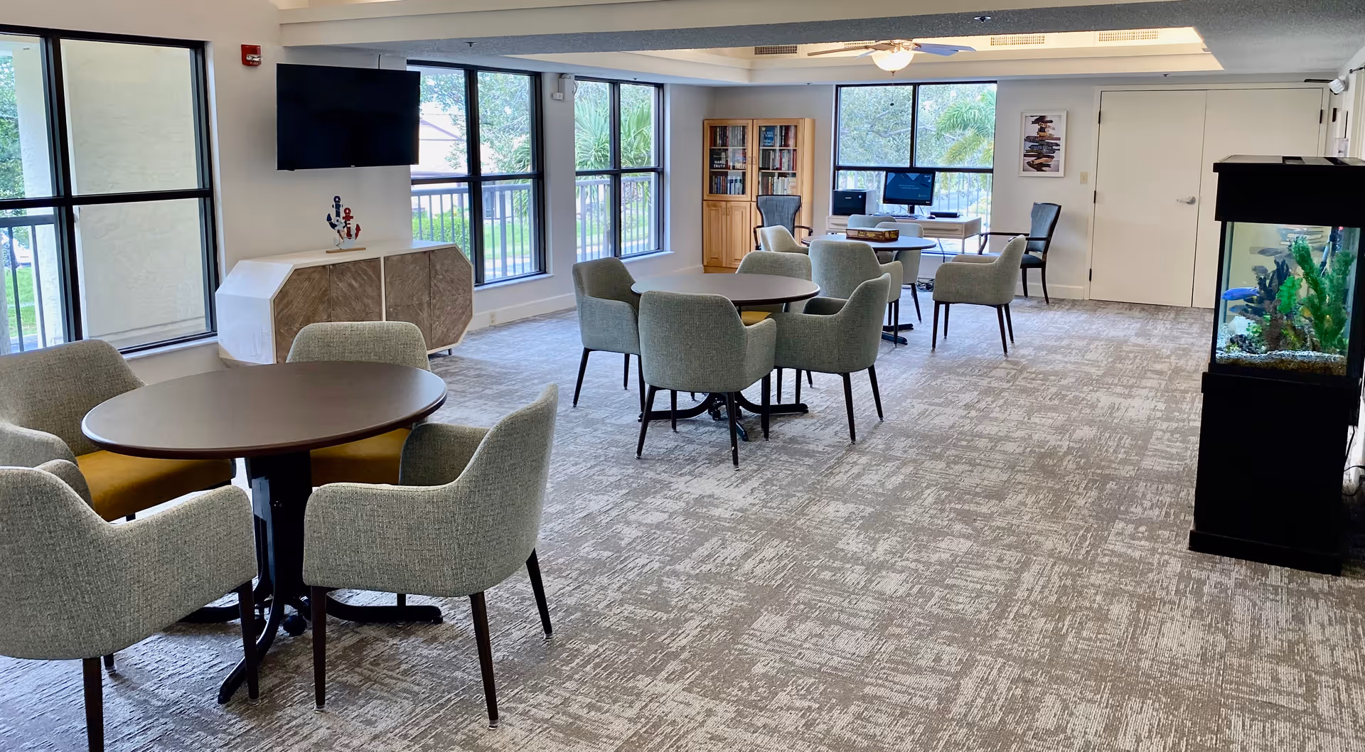 A bright and spacious common area in a senior living facility with multiple round tables surrounded by cushioned chairs. Large windows let in natural light, and there is a flat-screen TV mounted on the wall above a cabinet. A bookshelf and computer station are visible in the background, and a tall aquarium stands on the right side of the room.