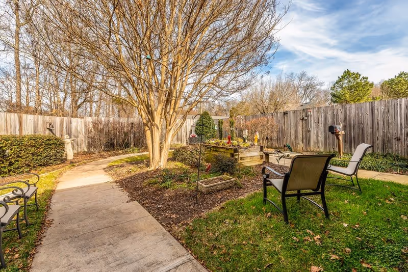 Outdoor garden area with a concrete pathway winding through it, surrounded by leafless trees and shrubs. There are several chairs placed on the grass near the path, and a wooden raised garden bed with bird feeders and decorations. A wooden fence encloses the area under a partly cloudy blue sky.