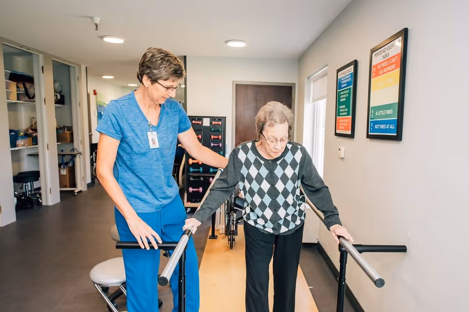 A healthcare worker in blue scrubs assists an elderly woman wearing a black and gray argyle sweater as she practices walking with parallel bars in a rehabilitation center hallway.