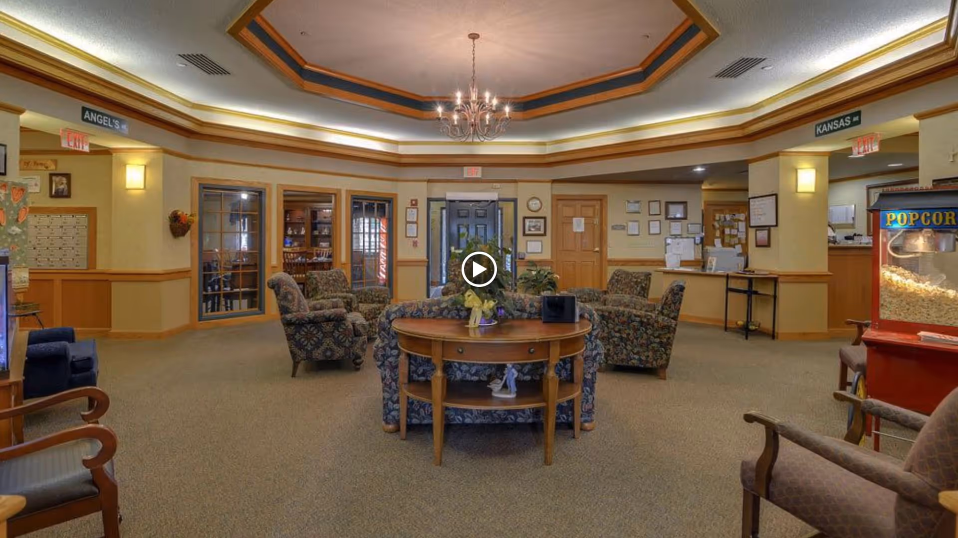 A spacious senior living facility common area with patterned armchairs arranged around a wooden table with decorative items. The room has beige walls with wooden trim, a chandelier hanging from a recessed ceiling, and a popcorn machine on the right side. There are doors and windows leading to other rooms, and signs labeled 'ANGELS' and 'KANSAS' above doorways.