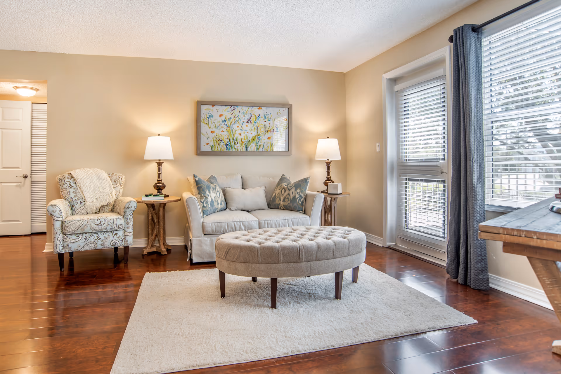 A cozy living room with a beige loveseat adorned with decorative pillows, a patterned armchair with a throw blanket, two wooden side tables each holding a lamp, a large oval tufted ottoman on a white rug, and a window with blinds and gray curtains letting in natural light.