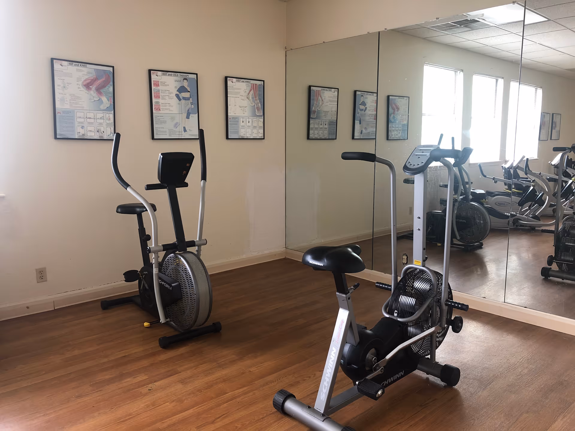 A small exercise room with two stationary exercise bikes on a wooden floor. The room has a large wall mirror reflecting the equipment and three framed anatomical posters on the wall. Natural light comes through the windows on the right side.
