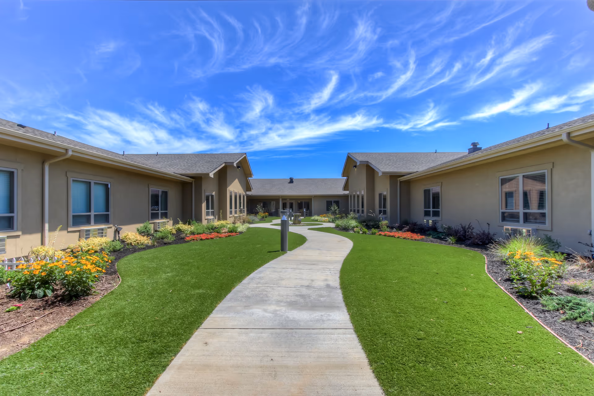 A landscaped courtyard with a winding concrete walkway between single-story assisted living buildings under a bright blue sky.