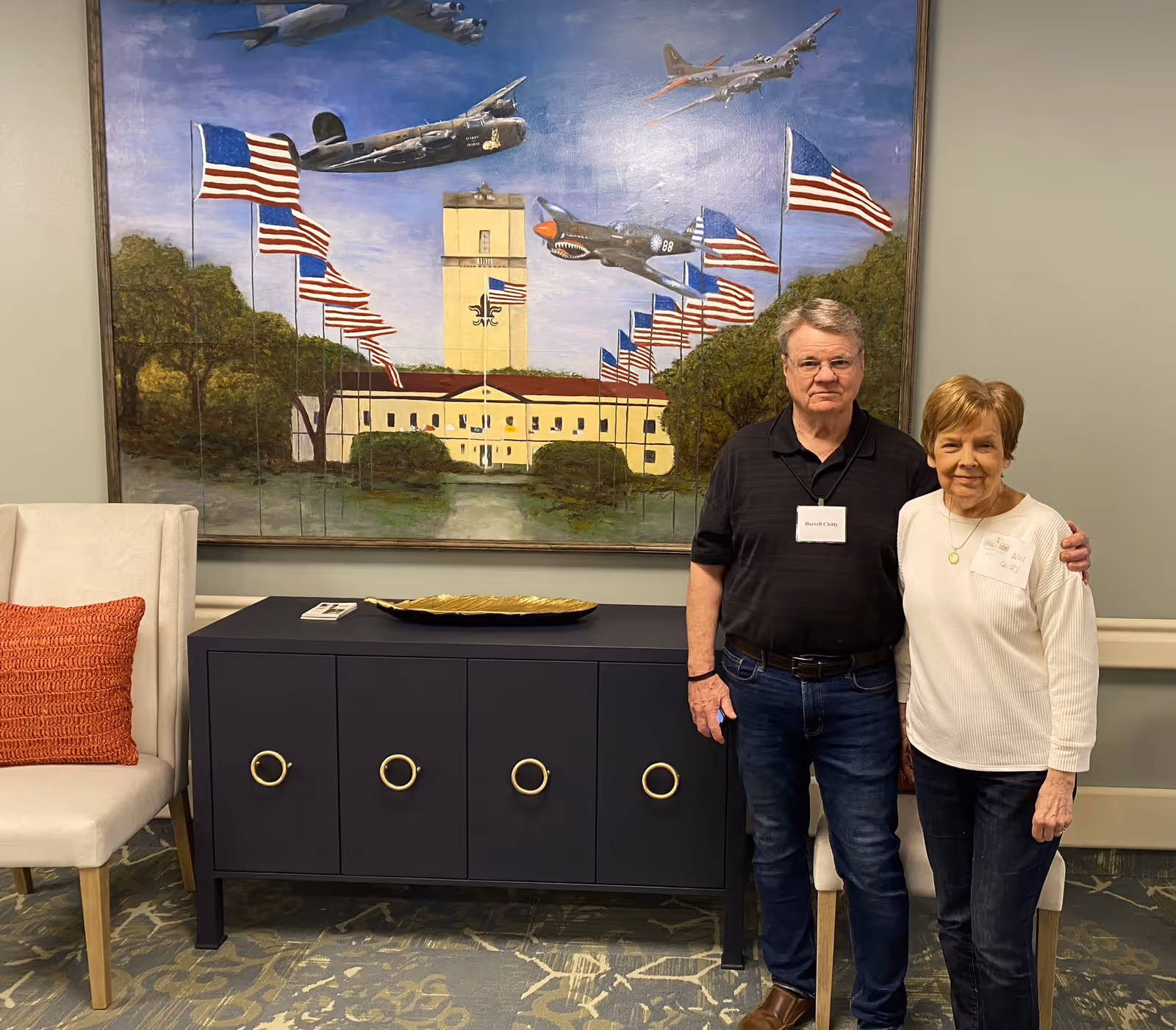 An elderly man and woman standing side by side in a room with a large painting behind them depicting American flags, trees, a building, and vintage airplanes flying in the sky. There is a black cabinet with gold ring handles and a decorative gold tray on top, and a white chair with an orange cushion to the left.