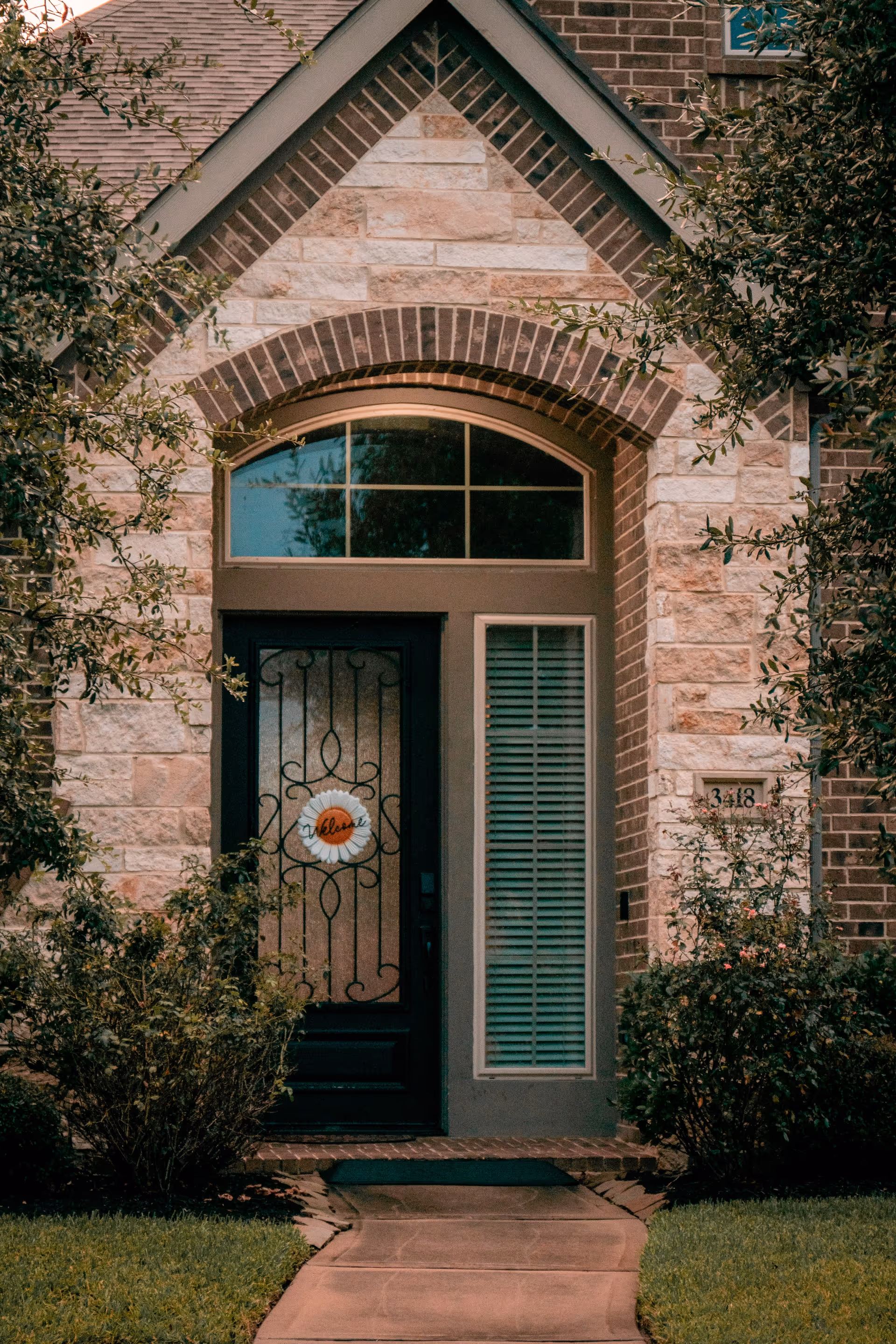 Front entrance of a house with a black door featuring decorative ironwork and a 'welcome' wreath. The door is framed by a large arched window above and a tall window with blinds to the right. The exterior is made of light-colored stone and brick, with bushes and greenery on either side of the walkway leading to the door.