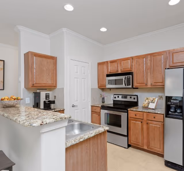 Modern kitchen with wooden cabinets, granite countertops, stainless steel appliances including a refrigerator, microwave, and oven. A sink is integrated into the countertop, and a coffee maker and a bowl of fruit are visible on the counter.