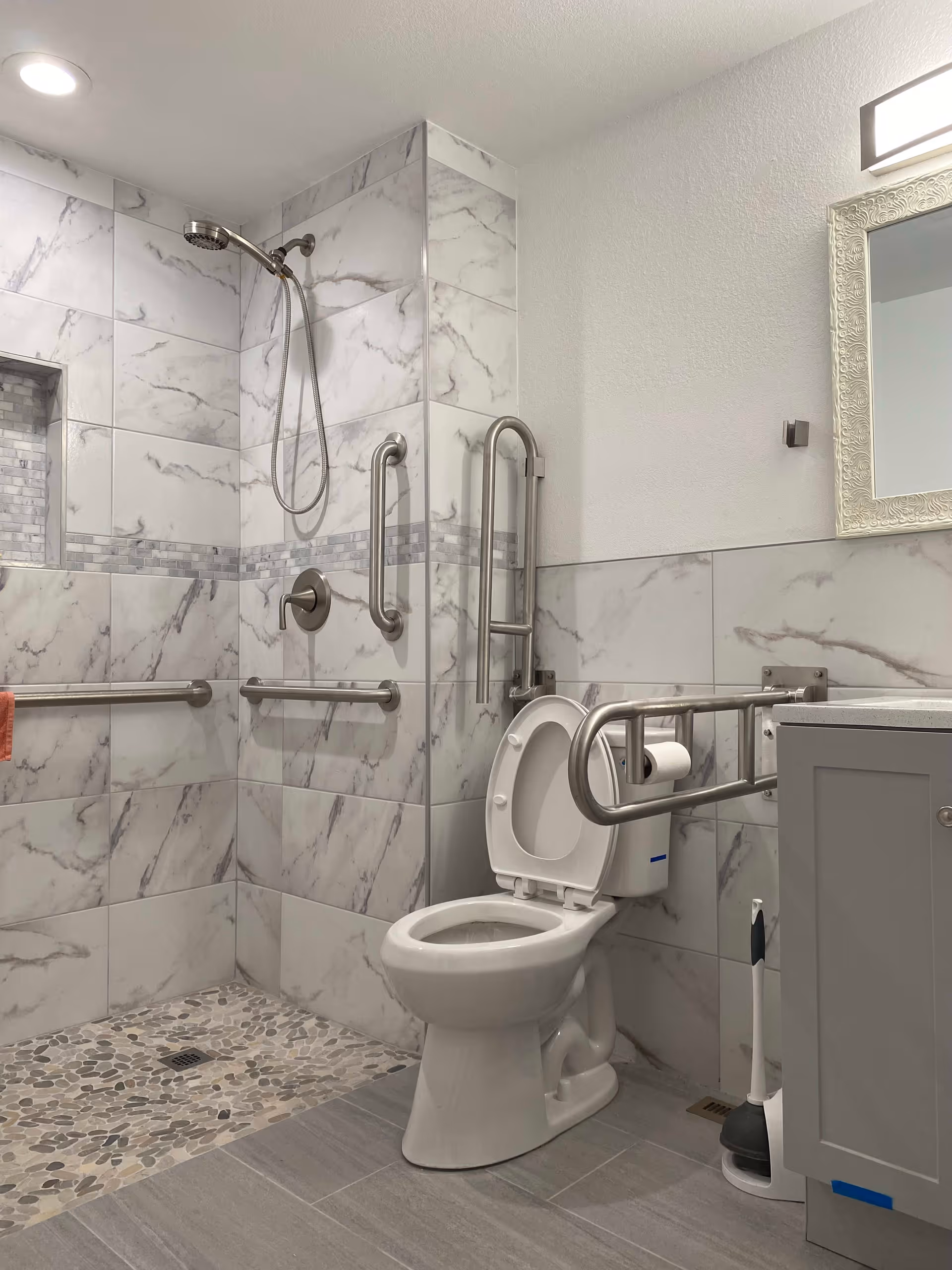 A modern bathroom with a walk-in shower featuring a handheld showerhead and multiple stainless steel grab bars. The walls are tiled with white and gray marble-patterned tiles, and the shower floor has pebble-style tiles. There is a white toilet with an open lid and a stainless steel grab bar with a toilet paper holder attached. A gray vanity with a white countertop and a decorative framed mirror above it are visible on the right side. A plunger is placed on the floor next to the vanity.