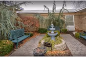 Outdoor courtyard area with a central three-tiered water fountain surrounded by greenery and plants. There are two green benches on either side of the paved walkway leading to the fountain. The building exterior with windows is visible in the background under an overcast sky.