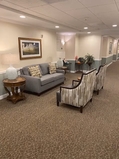 A cozy seating area in a senior living facility hallway featuring a gray sofa with patterned cushions, two striped armchairs, two round wooden side tables with lamps, a framed landscape picture on the wall, and a small table with plants and decorative items in the background.