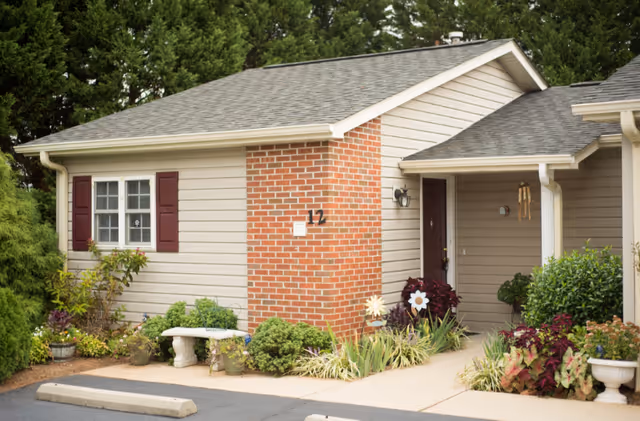 Exterior view of a single-story residential building with beige siding and a brick chimney. The building has a small covered entrance with a dark red door, surrounded by well-maintained shrubs and plants. There is a white bench near the window with maroon shutters, and the building number 12 is visible on the brick chimney. Tall green trees are in the background.