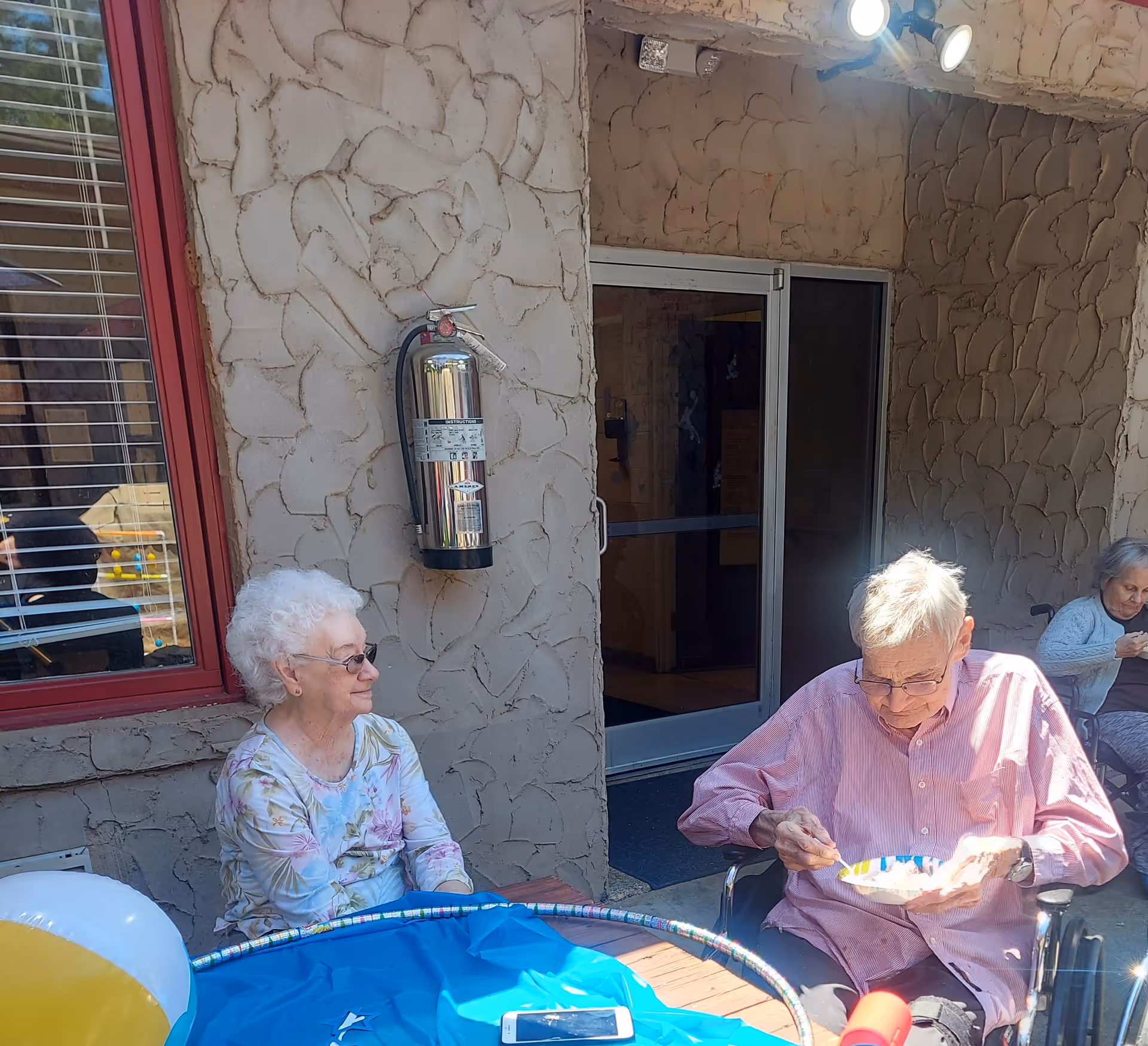 Two elderly residents sit at an outdoor patio table with party decorations while a fire extinguisher is mounted on the stucco wall behind them.