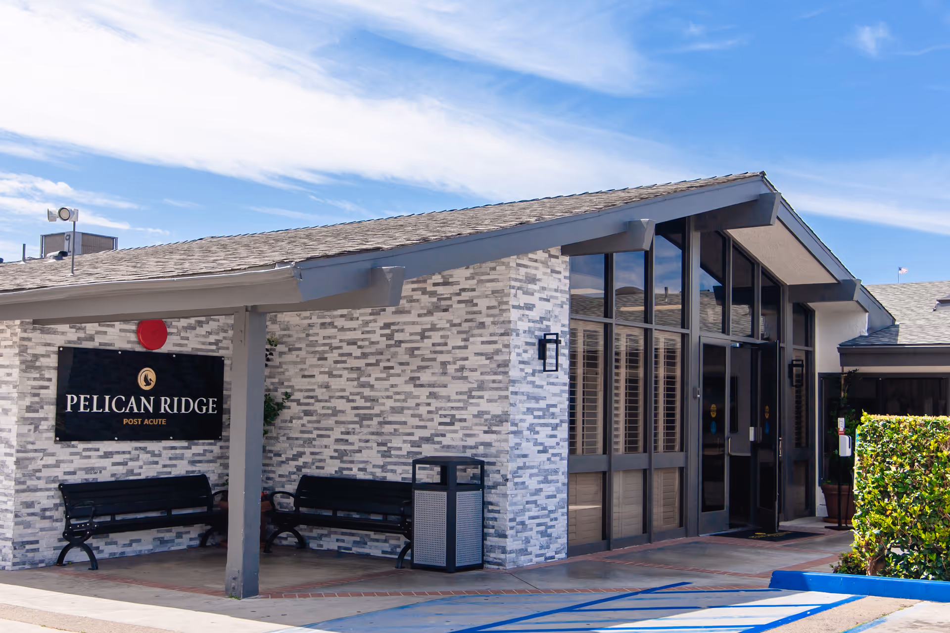 Exterior view of the Pelican Ridge Post-Acute facility showing the entrance with large windows, a covered walkway, two black benches, a trash can, and a sign with the facility name on a gray brick wall under a blue sky with some clouds.