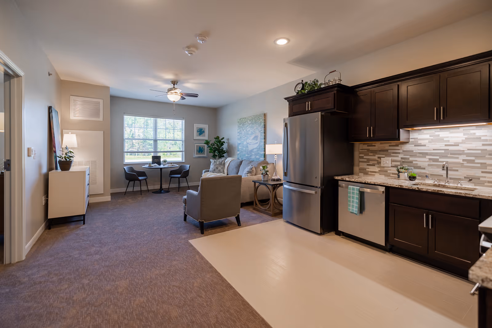 Interior view of a senior living facility apartment featuring a modern kitchen with dark wood cabinets, stainless steel refrigerator and dishwasher, and a tiled backsplash. Adjacent to the kitchen is a living area with a gray armchair, a sofa, a side table with a lamp, and wall art. A small dining area with a round table and two chairs is positioned near a window with blinds, allowing natural light to fill the room.