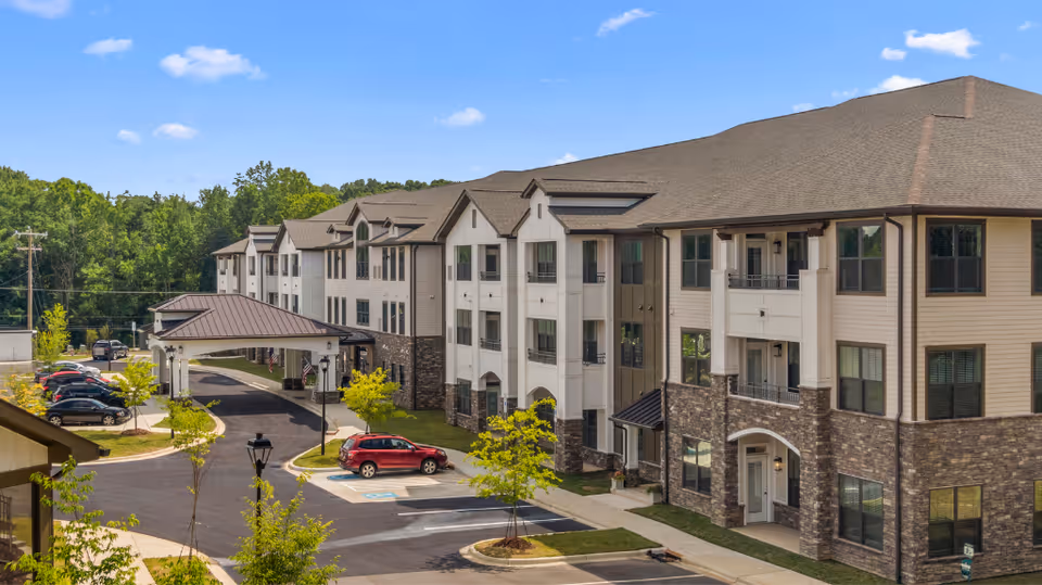 Exterior view of Ambriant at Austin Village senior living facility showing a three-story building with a covered entrance, multiple windows, and a parking lot with several cars. The building has a combination of stone and light-colored siding, surrounded by trees and greenery under a blue sky with some clouds.
