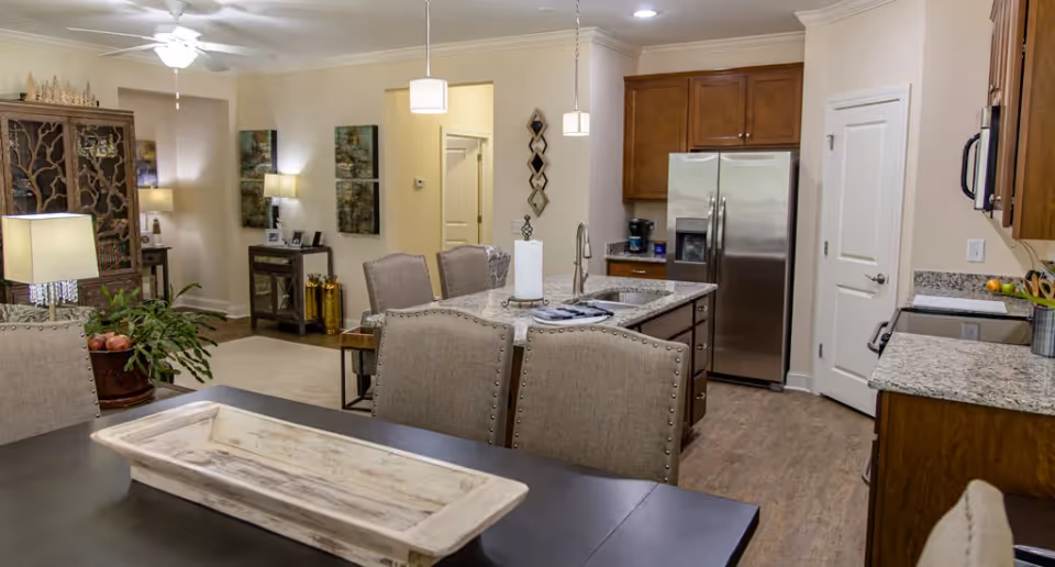 Open-plan kitchen and dining area with a central island, stainless steel refrigerator, granite countertops, and upholstered dining chairs with a living room visible in the background.