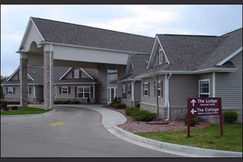 Exterior view of a senior living facility building with a covered entrance and multiple windows. There is a paved driveway leading to the entrance, and a sign on the lawn indicating directions to 'The Lodge' and 'The Cottage'. The sky is partly cloudy.