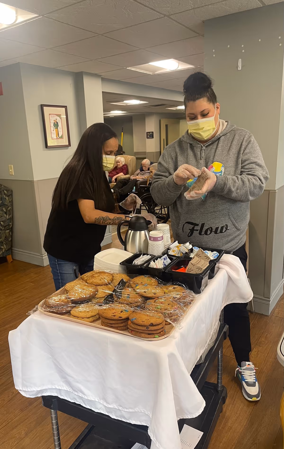Two women wearing face masks and gloves preparing a snack cart with cookies, coffee, and condiments in a senior living facility. In the background, elderly residents are seated in chairs and wheelchairs in a common area.