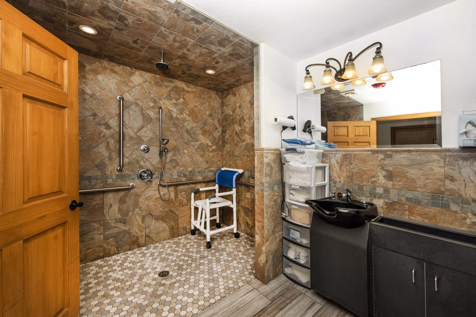 Accessible bathroom with a walk-in shower featuring brown stone tiles on the walls and ceiling, a white shower chair with a blue backrest, and multiple grab bars. The bathroom also includes a black sink with a cabinet underneath, a large mirror with four light fixtures above, and a plastic drawer organizer holding towels and supplies. A wooden door is partially open to the left.