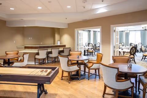 A spacious common area in a retirement community featuring several round wooden tables with cushioned chairs arranged around them. In the foreground, there is a shuffleboard table. The room has light-colored walls and wooden flooring. Through two open doorways, additional seating areas with more tables and chairs are visible, with large windows allowing natural light to enter.
