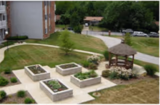 Outdoor garden area with raised rectangular flower beds, a wooden gazebo, and a paved walkway. The garden is adjacent to a multi-story brick building with green trees and parked cars in the background.