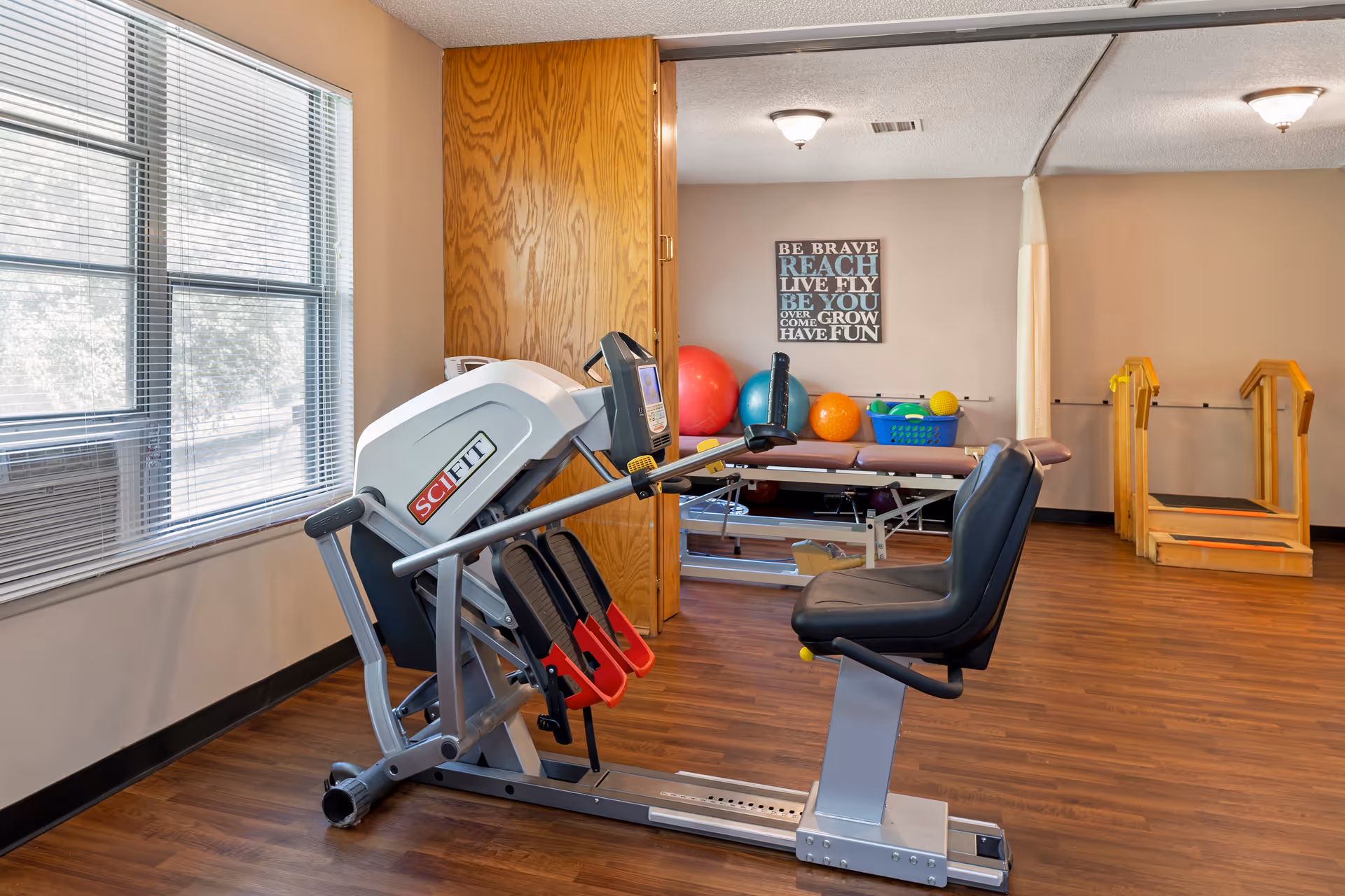 A rehabilitation room with exercise equipment including a SciFit machine, therapy balls, a treatment table, and wooden stairs for physical therapy. The room has wood flooring, beige walls, and a large window with blinds letting in natural light. A motivational wall sign reads 'Be brave, reach, live, fly, be you, overcome, grow, have fun.'