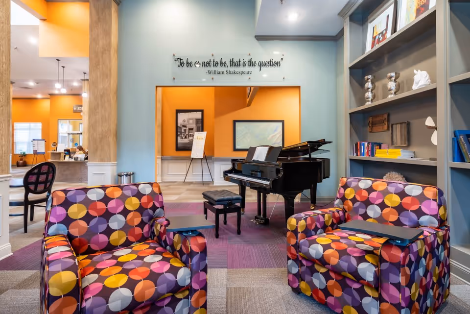 Bright common lounge with colorful patterned armchairs, a grand piano, bookshelf and a reception area in the background.