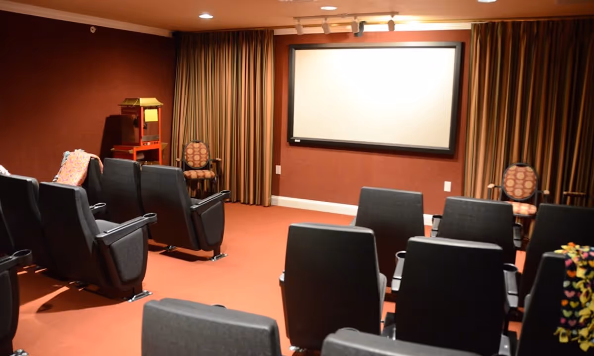 A small theater room with rows of black cushioned seats facing a large blank projection screen mounted on a red wall. The room has red carpeting and striped curtains on the windows. There is a popcorn machine and two patterned armchairs in the back corners of the room.