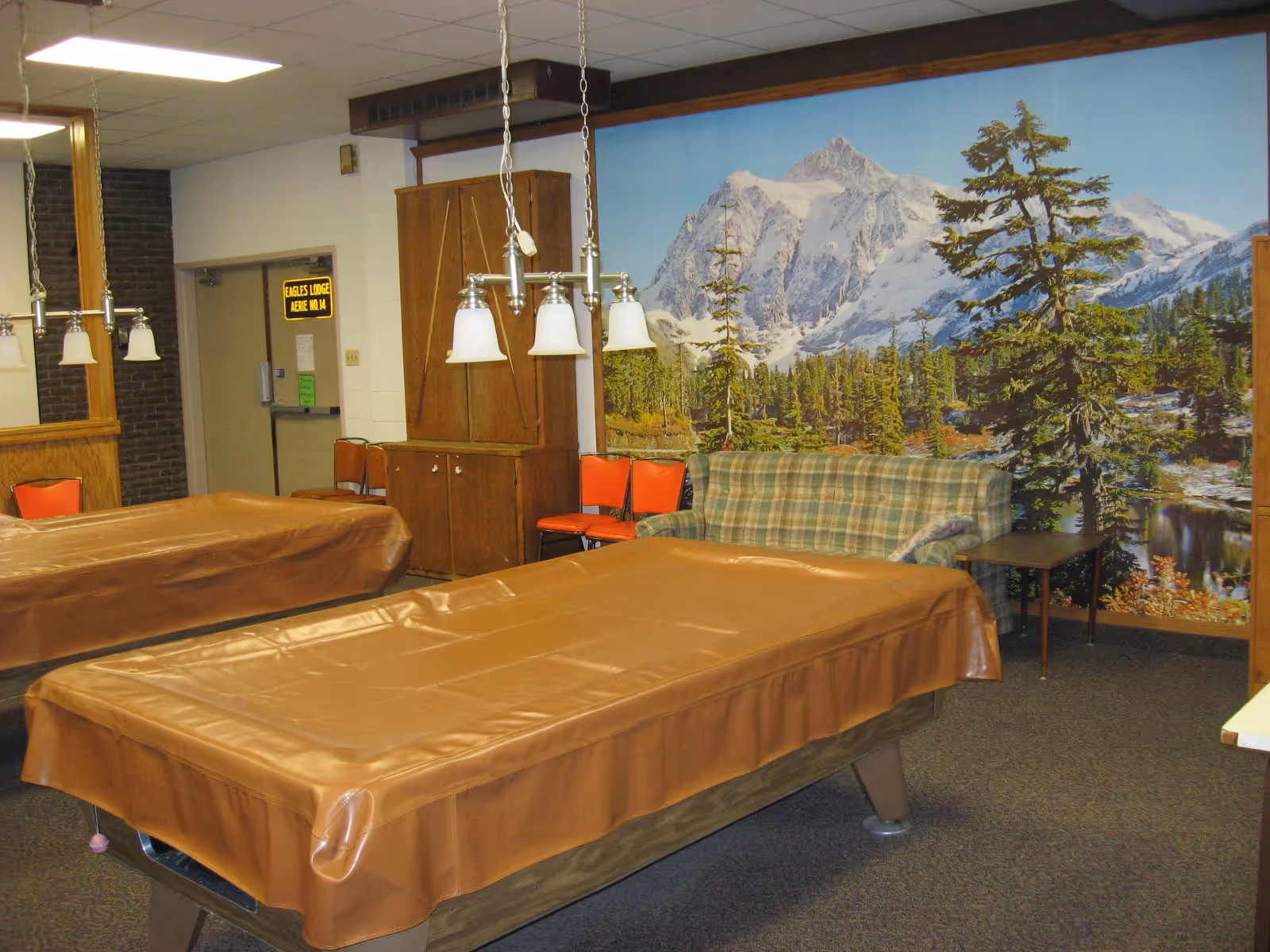 A recreational room with two pool tables covered in brown protective covers, a plaid couch, several orange chairs, wooden cabinets, and a large wall mural depicting a snowy mountain landscape with pine trees.