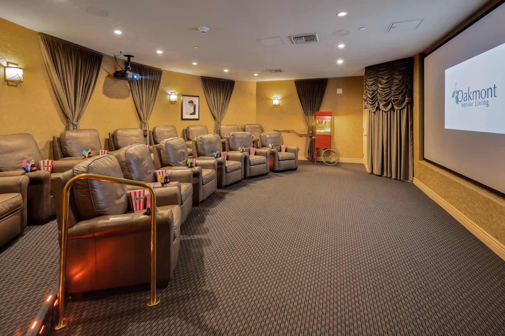 A cozy senior living facility theater room with two rows of comfortable leather recliners, each equipped with popcorn and candy. The room has warm lighting, curtains on the windows, a popcorn machine in the corner, and a large screen displaying the Oakmont Senior Living logo.