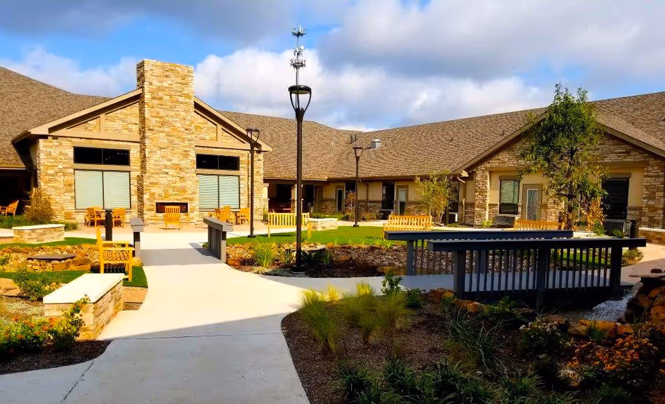 Outdoor courtyard area of Cedar Pointe Wellness And Health Center featuring a stone building with large windows, a central stone chimney, multiple wooden benches, landscaped garden beds, a small footbridge over a dry creek bed, and several lamp posts under a partly cloudy sky.
