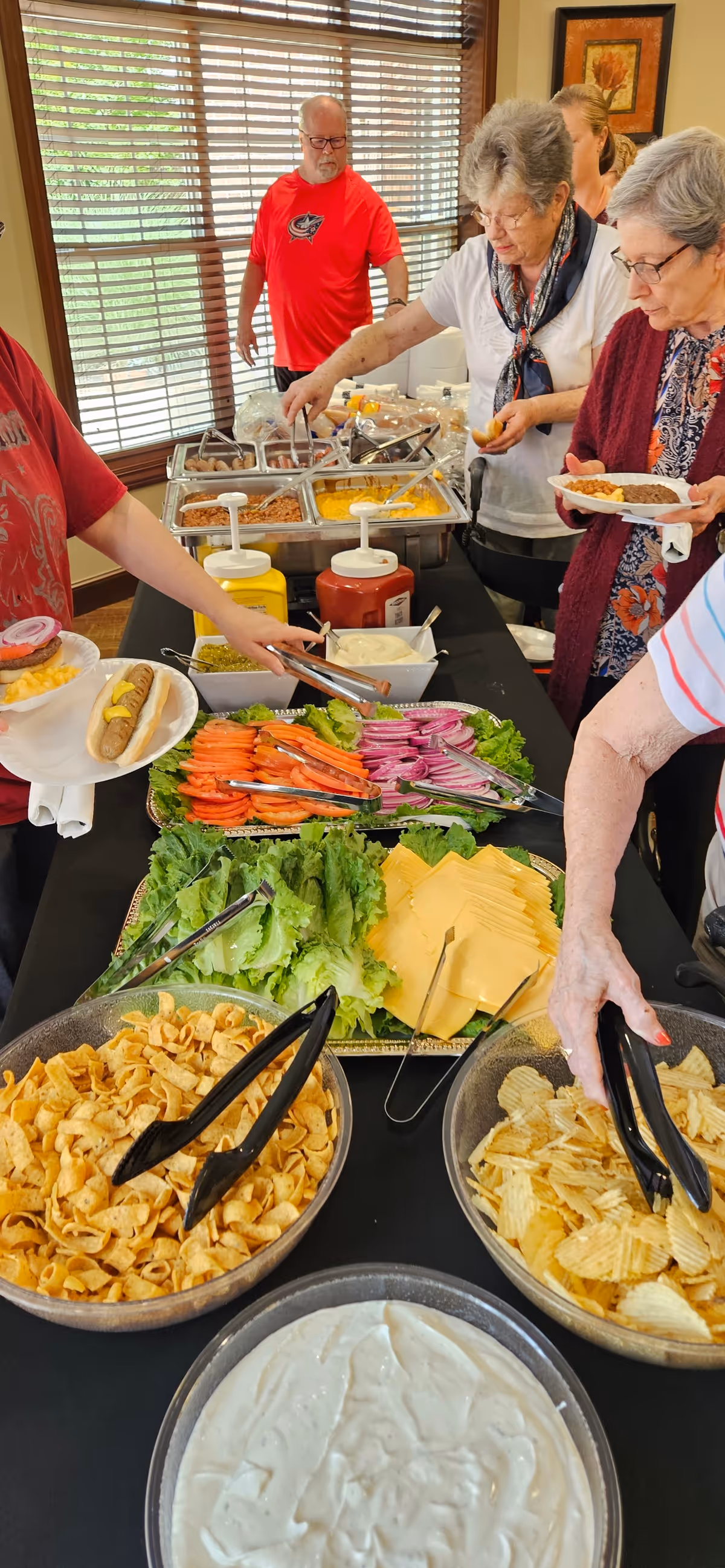 Several elderly people serving themselves food from a buffet table that includes chips, sliced cheese, lettuce, tomato, onions, condiments, and hot dogs. The setting appears to be indoors with large windows in the background.