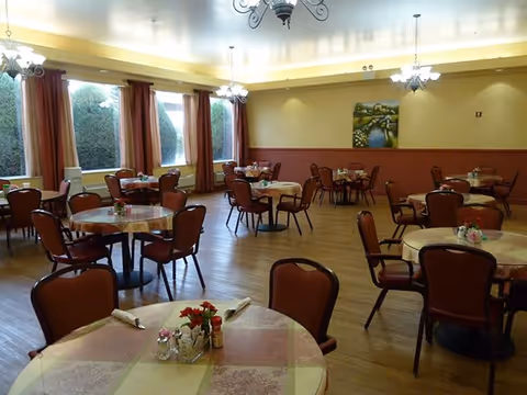 Spacious assisted-living dining room with round tables and chairs set for meals, chandeliers, and large windows with curtains.