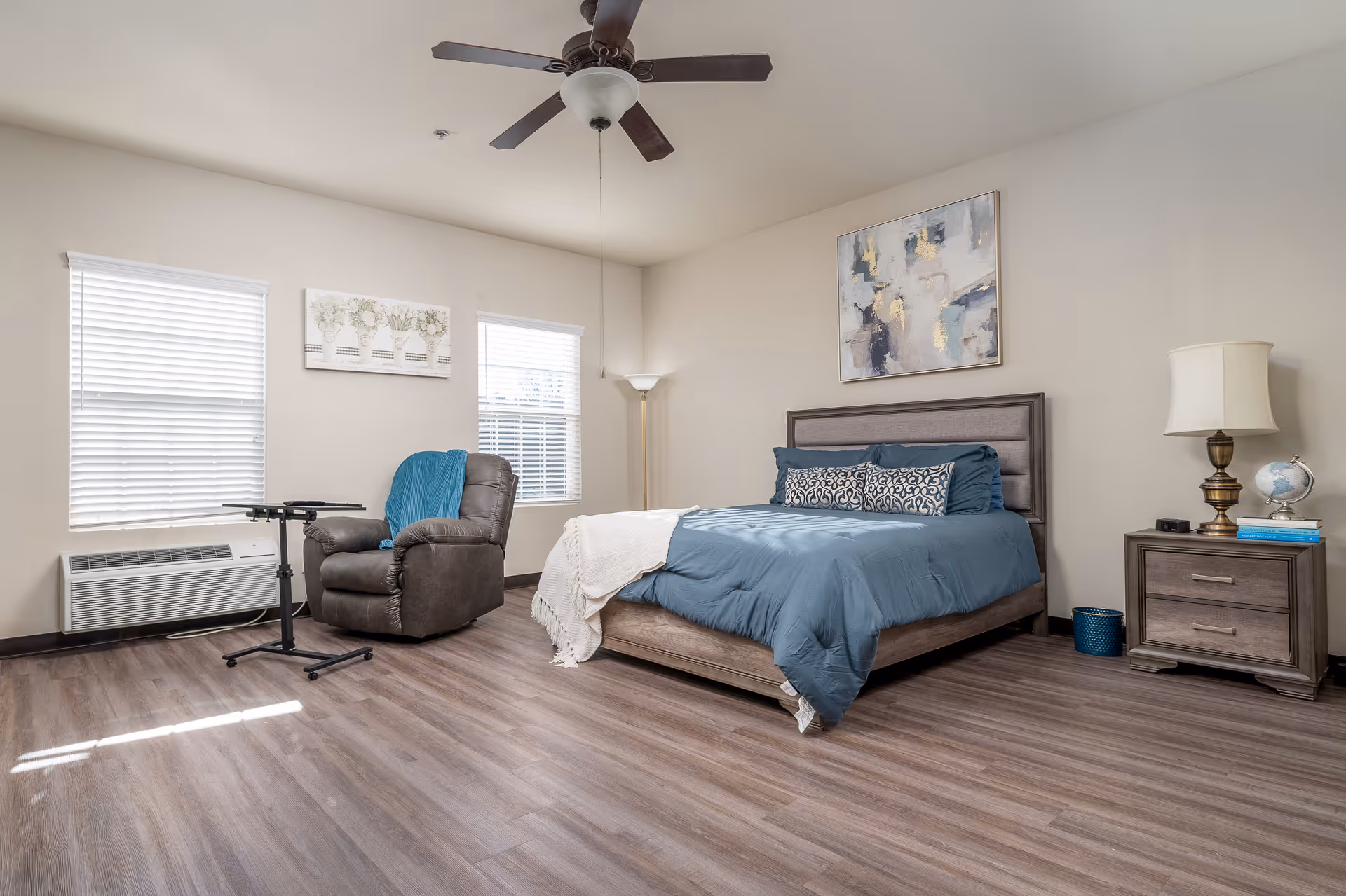 A spacious bedroom with a large bed dressed in blue bedding and decorative pillows. To the right of the bed is a wooden nightstand with a lamp, a globe, and some books. On the left side of the room, there is a comfortable recliner chair with a blue throw blanket and a small adjustable table in front of it. Two windows with white blinds allow natural light to fill the room. The walls are light-colored, and there is a ceiling fan overhead. A piece of abstract artwork hangs above the bed.